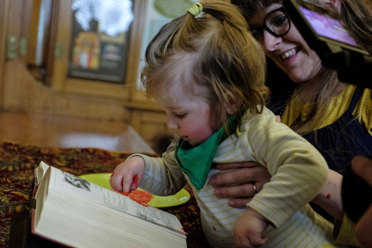 A baby, held by his mother, spreads paint on a book.